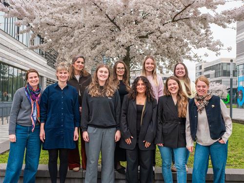 Eine Gruppe von zehn Frauen unterschiedlichen Alters steht lächelnd vor einem blühenden Kirschbaum auf einem modernen Hochschulcampus. Die Personen sind in zwei Reihen angeordnet und blicken in die Kamera, im Hintergrund sind Gebäude und Frühlingsblüten zu sehen.